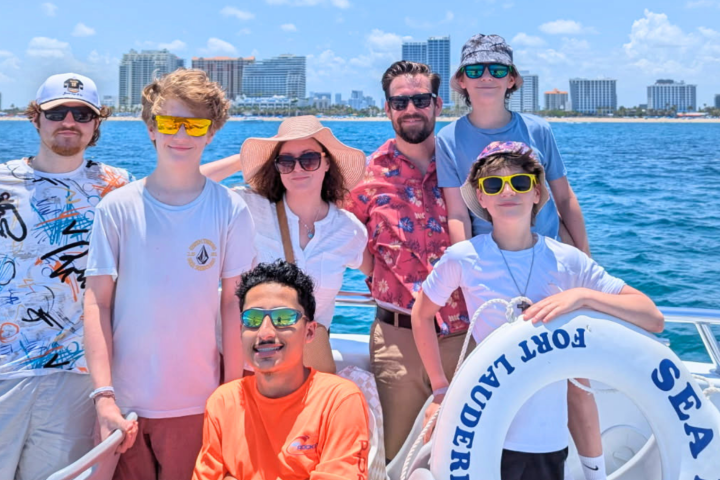 Group of people wearing sunglasses on a boat with cityscape in the background.