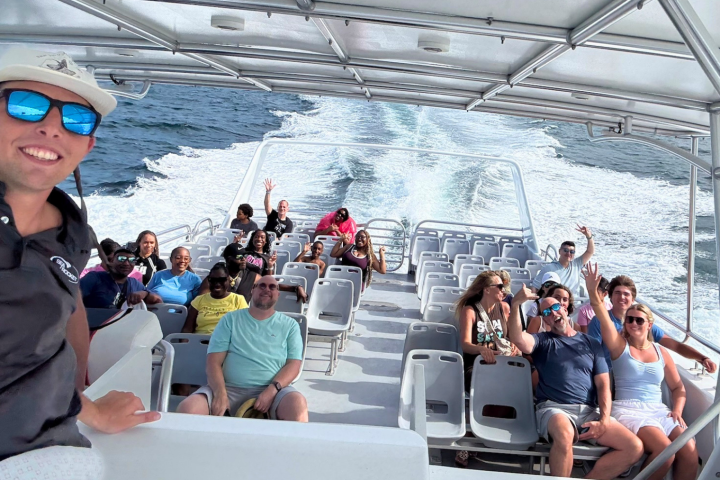 Group of people on a boat smiling and waving with the sea in the background.