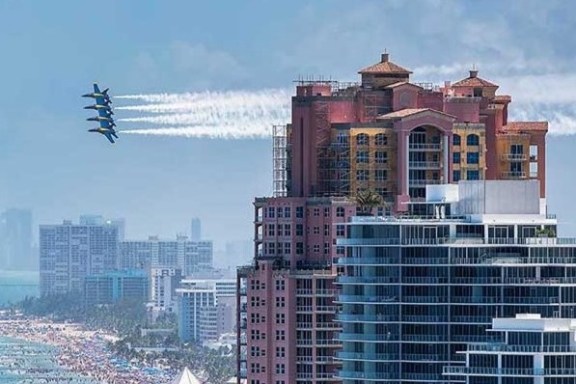 Jets flying by tall buildings near a crowded beach.
