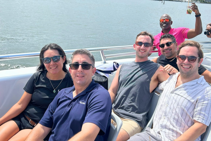 Group of people smiling on a boat, sunny day, water in background.