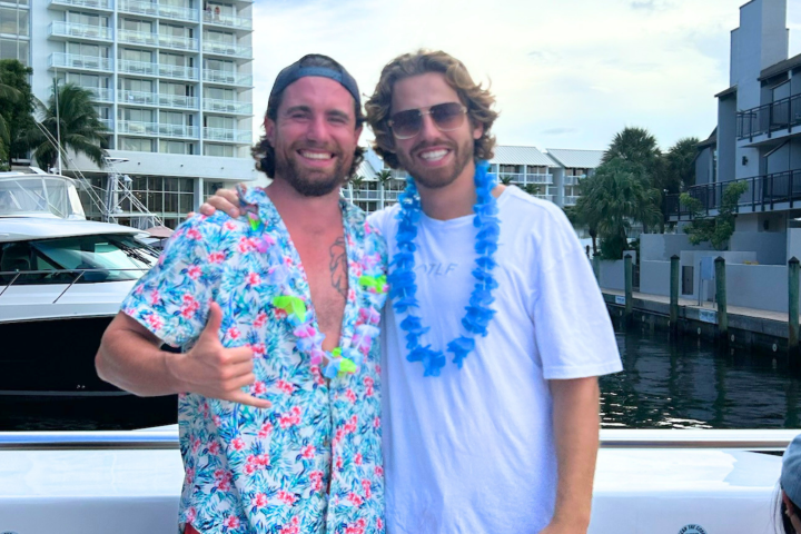 Two men in floral shirts smiling on a boat near a marina.