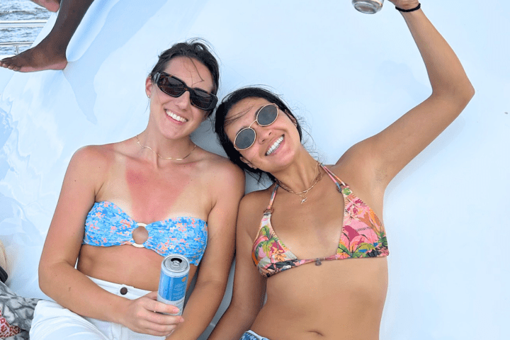 Two women in swimsuits smiling on a boat, holding drinks.