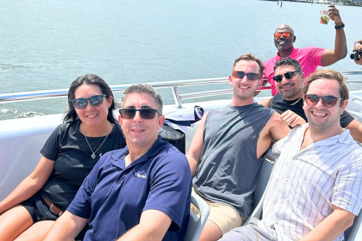 Group of six people smiling on a boat, wearing sunglasses, with water in the background.