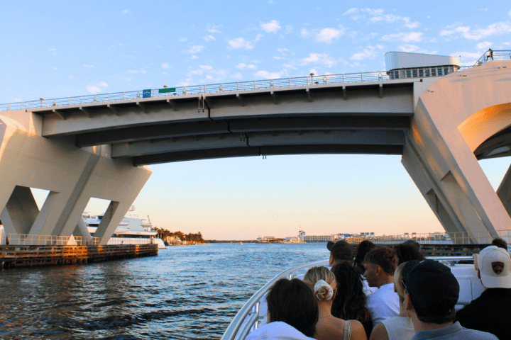 People on a boat passing under a large concrete bridge over a waterway.