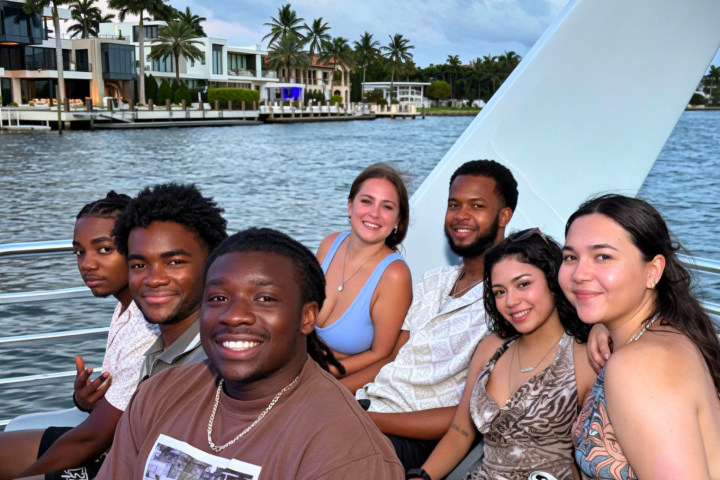 Group of seven people smiling on a boat with waterfront houses in the background.