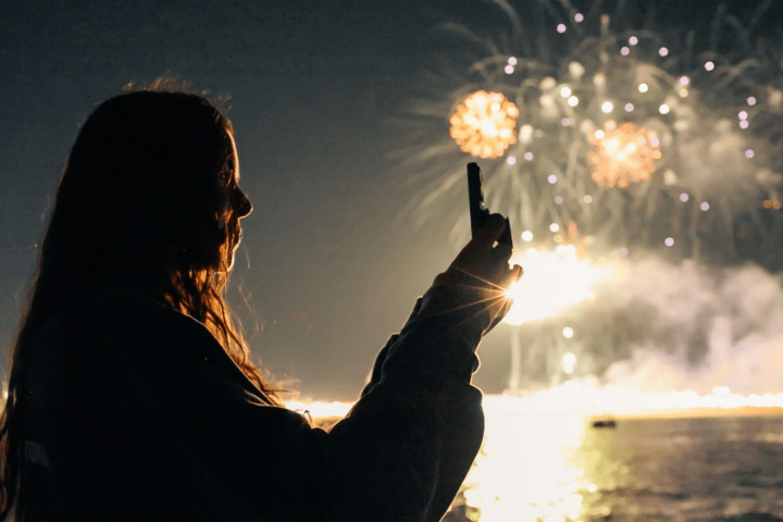 Silhouette of a person taking a photo of fireworks over water at night.