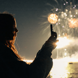 Silhouette of person taking photo of fireworks over water at night.