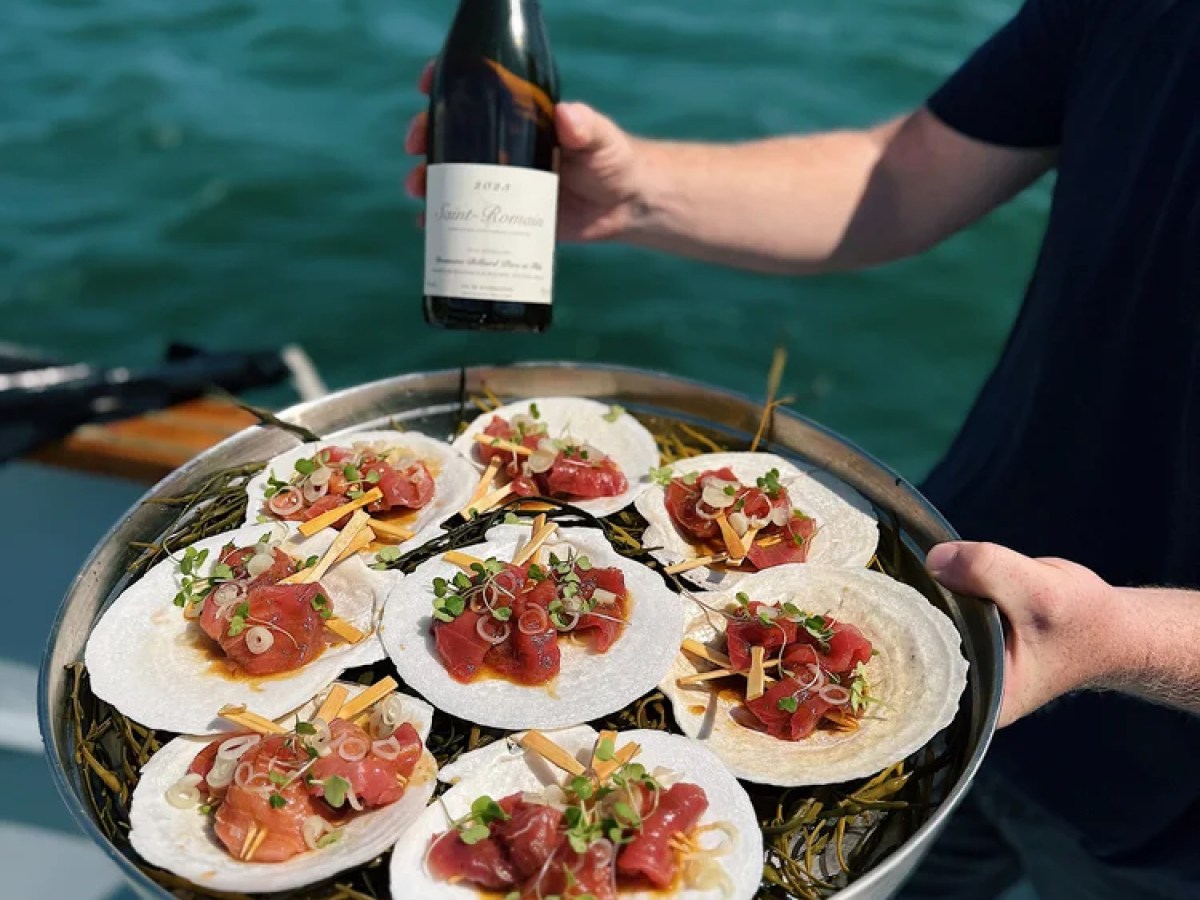 Person holds tray of tuna scallops and wine on a boat by the ocean.