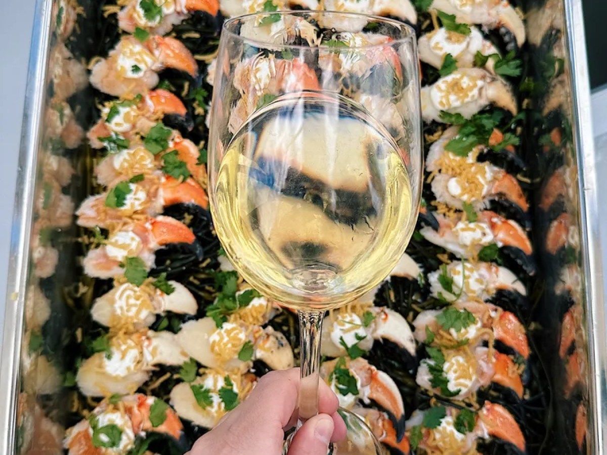 Hand holding a glass of white wine over a tray of assorted sushi.