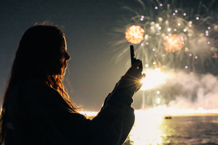 Silhouette of a person taking a photo of fireworks over water at night.
