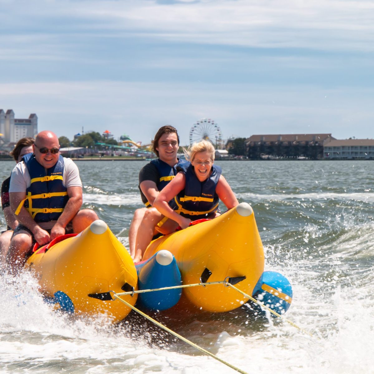 Four people riding a banana boat on the ocean with a city skyline in the background.