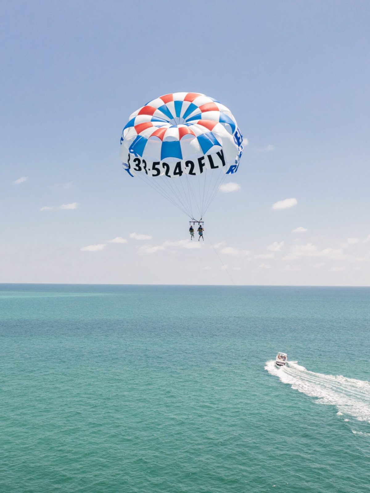 Parasailing duo under a red, white, and blue parachute above the ocean, boat trailing below.