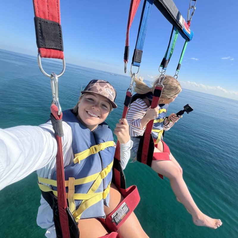 Two people parasailing over the ocean on a sunny day, wearing life vests and harnesses.