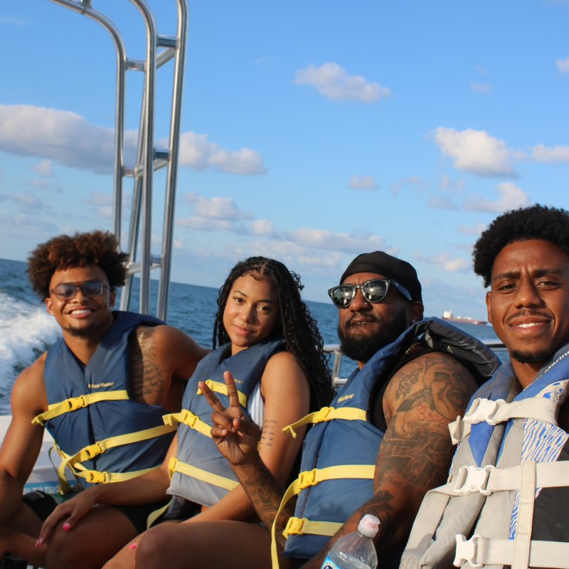 Four people in life vests sitting on a boat, smiling, with blue sky and water in the background.